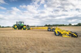 SWIFTERDISC XE_MEGA folded to a 3 m transport width, towed by a John Deere track tractor between fields